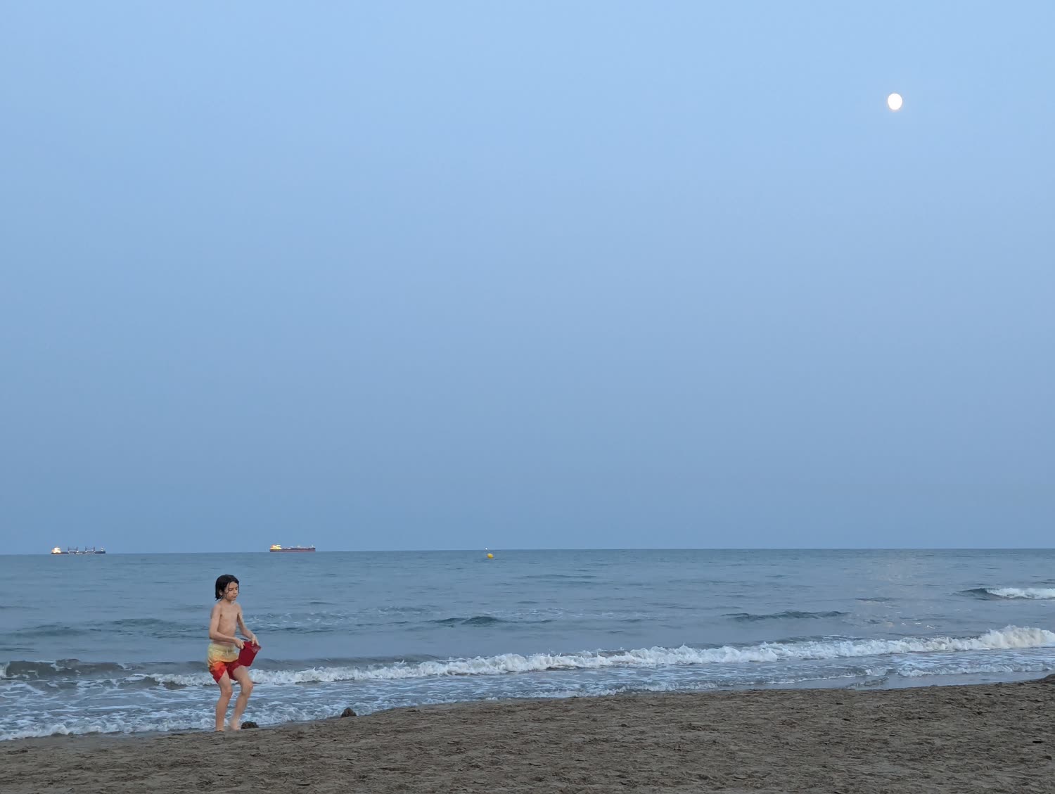 Un niño con un cubo en la orilla, al atardecer, con la luna.